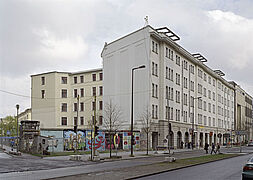 Blick über eine Straße auf ein fünfgeschossiges Haus an dessen linker Flanke ein Stück buntbemalter Berliner Mauer anschließt.