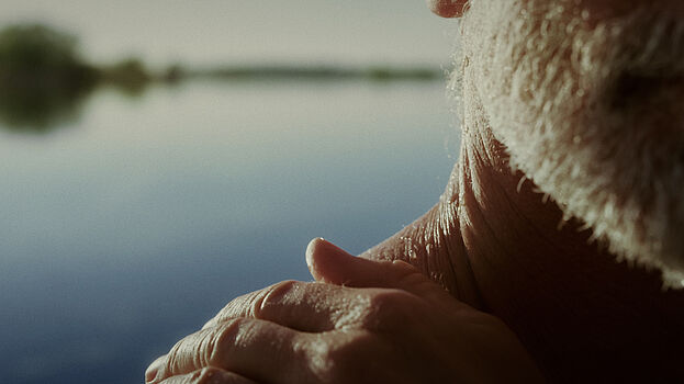 Ein Ausschnitt eines Mannes, der seine Schulter berührt. Im Hintergrund ist ein See zu sehen.