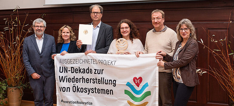 Steffi Lemke hält mit fünf anderen ein Banner mit der Aufschrift "Ausgezeichnetes Projekt: UN-Dekade zur Wiederherstellung von Ökosystemen"
